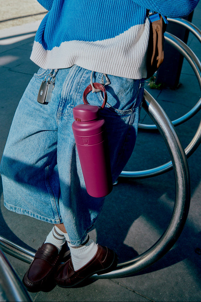 Person holding a mulberry water bottle with a blue sweater and jeans, standing next to a bicycle rack.