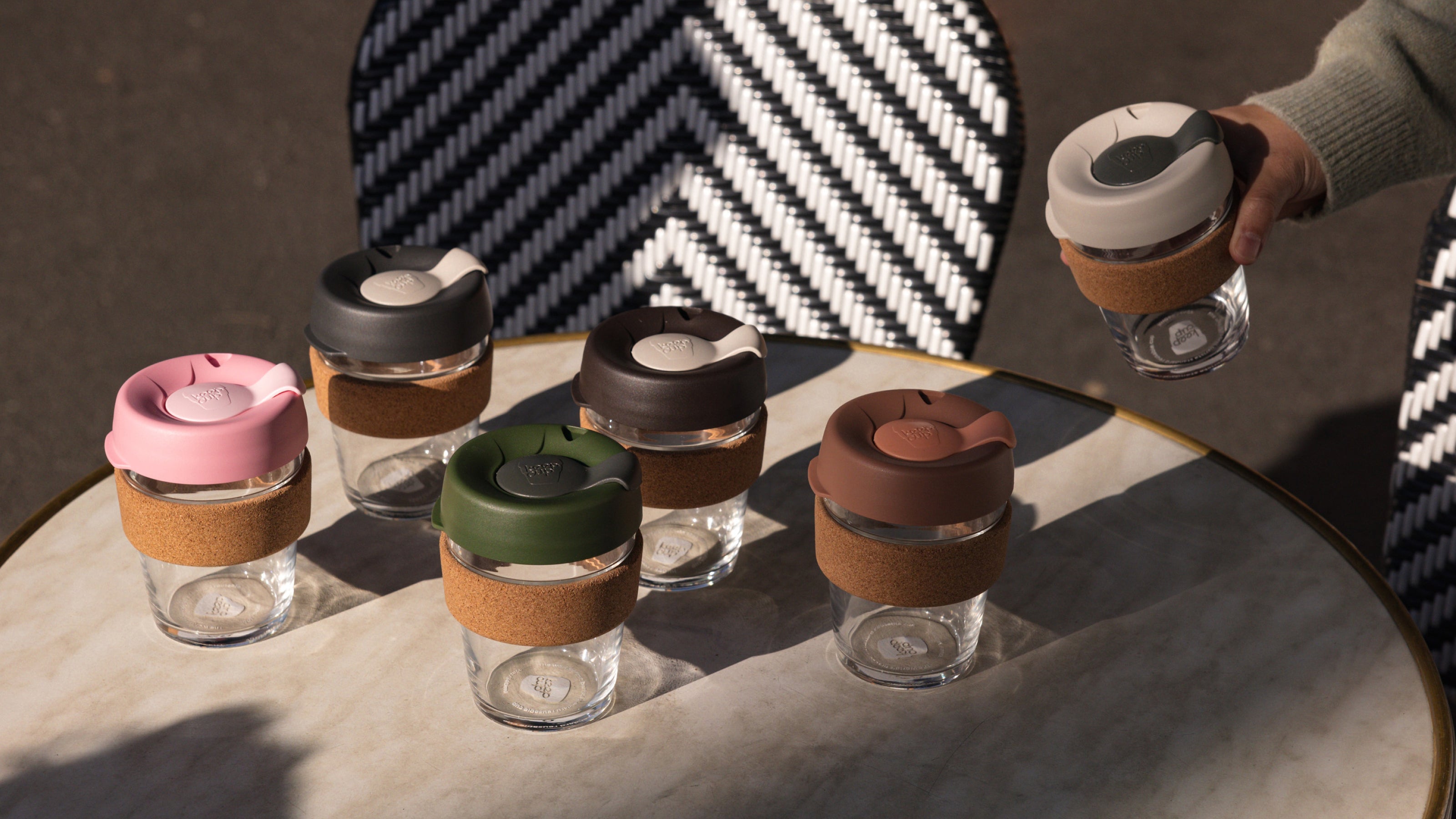 Colourful reusable glass coffee cups with cork bands and silicone lids in pink, green, black, brown and grey arranged on wooden table