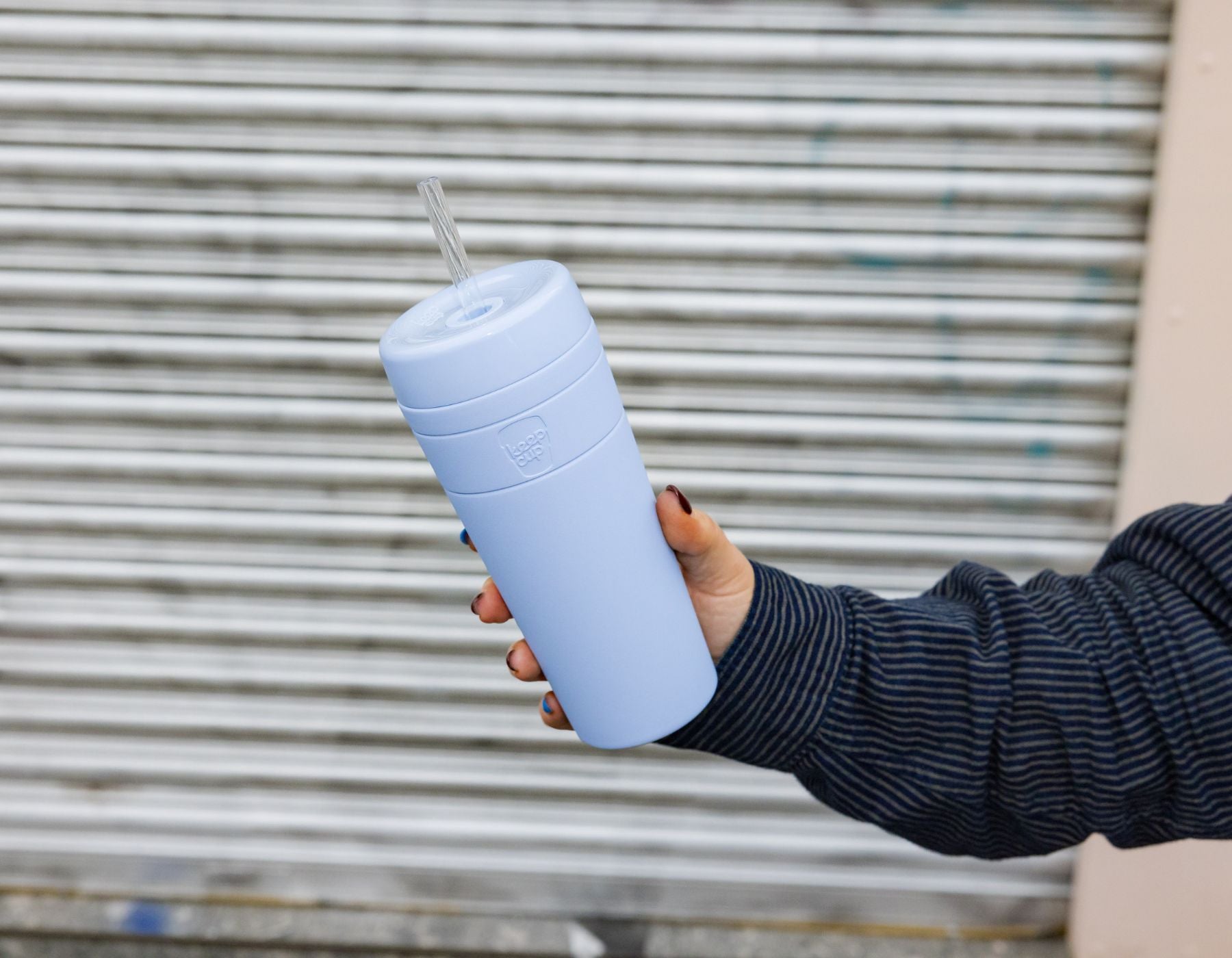 Light blue reusable tumbler with straw held in hand, wearing navy striped sleeve against metal shutters