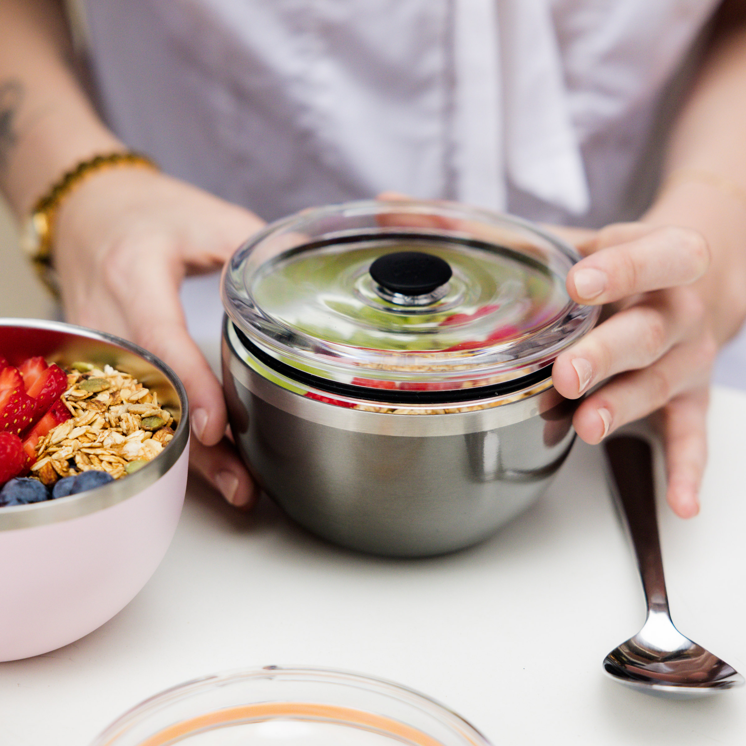 Stainless steel insulated food container with clear lid being held by hands, with breakfast bowl containing granola and berries nearby