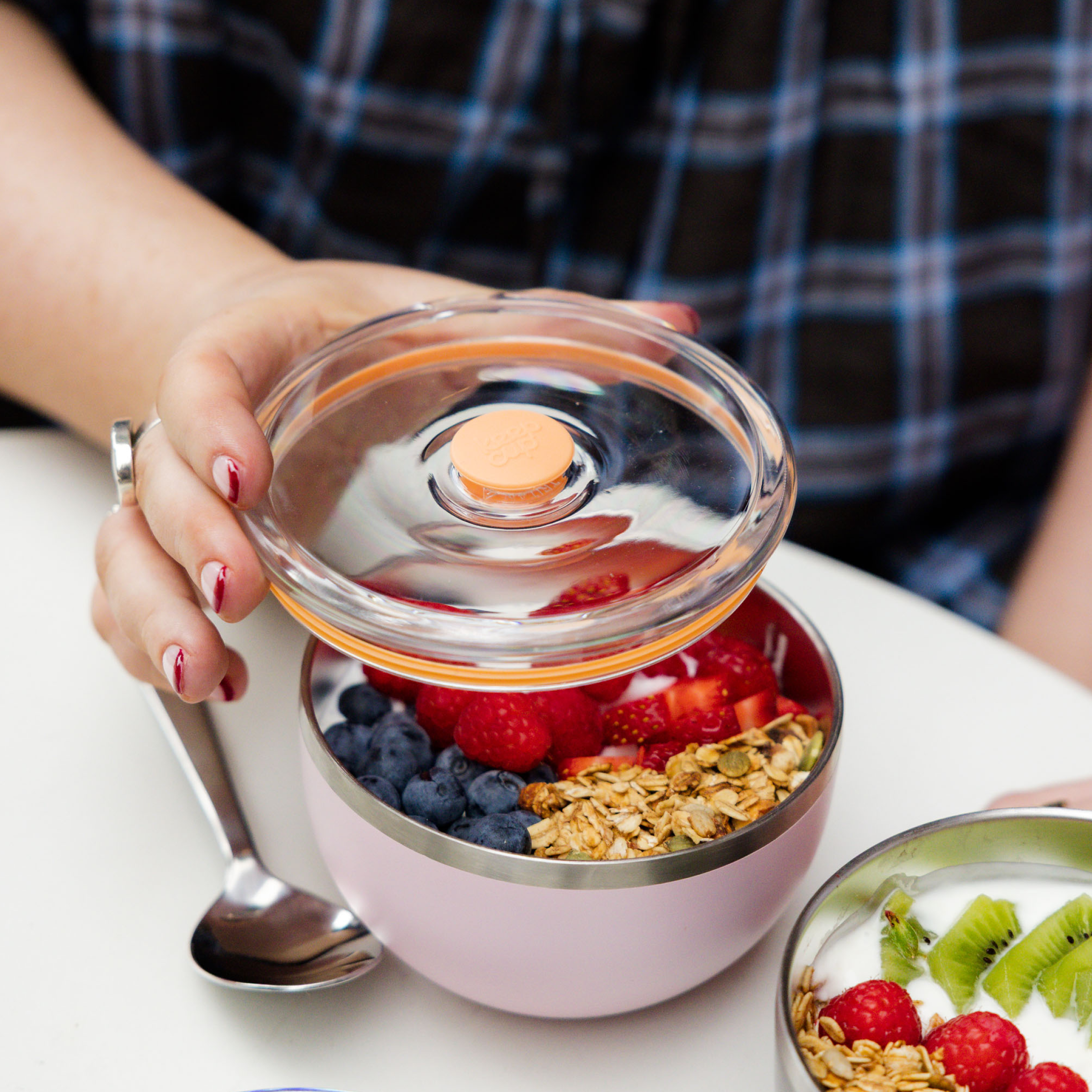 A hand with red nail polish lifts the clear plastic lid off a KeepCup Go High Go Low in Pashmak Pink, revealing a breakfast bowl filled with blueberries, raspberries, strawberries and granola, with a metal spoon resting against the cup on a white table.