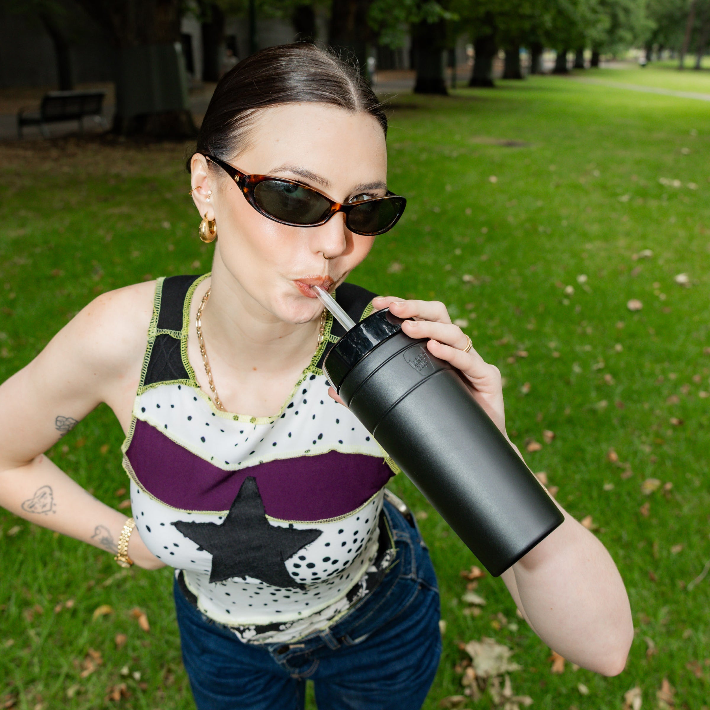 A woman wearing sunglasses and a patterned top drinks from a black KeepCup Commuter Cold Cup XL 20oz using the straw, photographed at a slight low angle in a sunlit park setting.