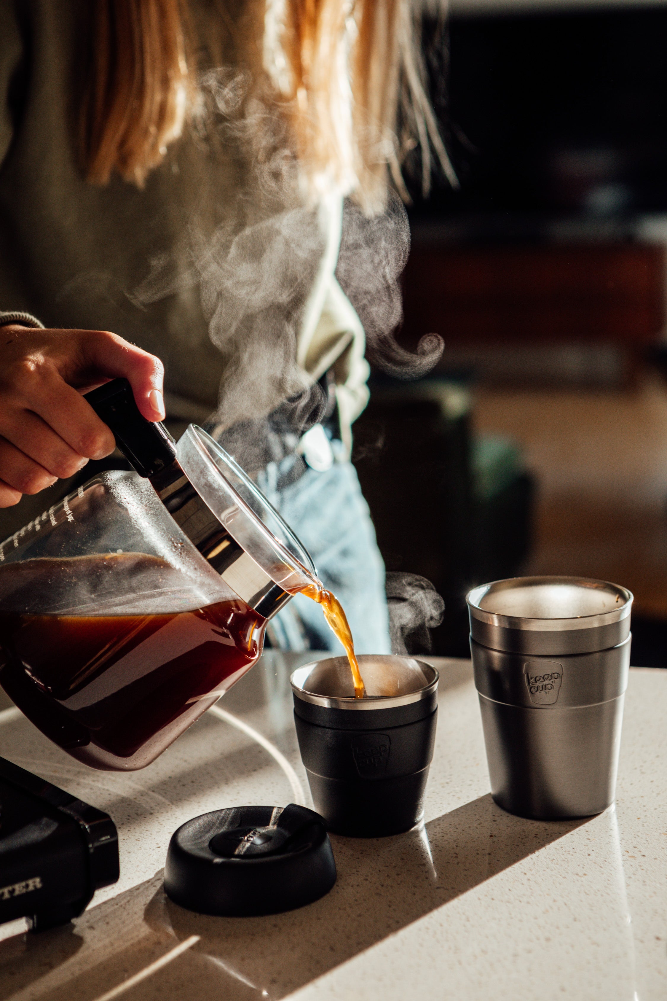 Person pouring coffee from a French press into a cup with steam rising, set against a blurred indoor background.