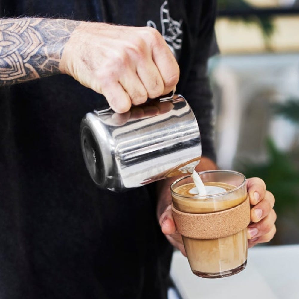 A barista with tattooed arms pours steamed milk from a stainless steel pitcher into a KeepCup Brew Cork M 12oz in Moonlight, creating latte art in the glass cup with its natural cork band.