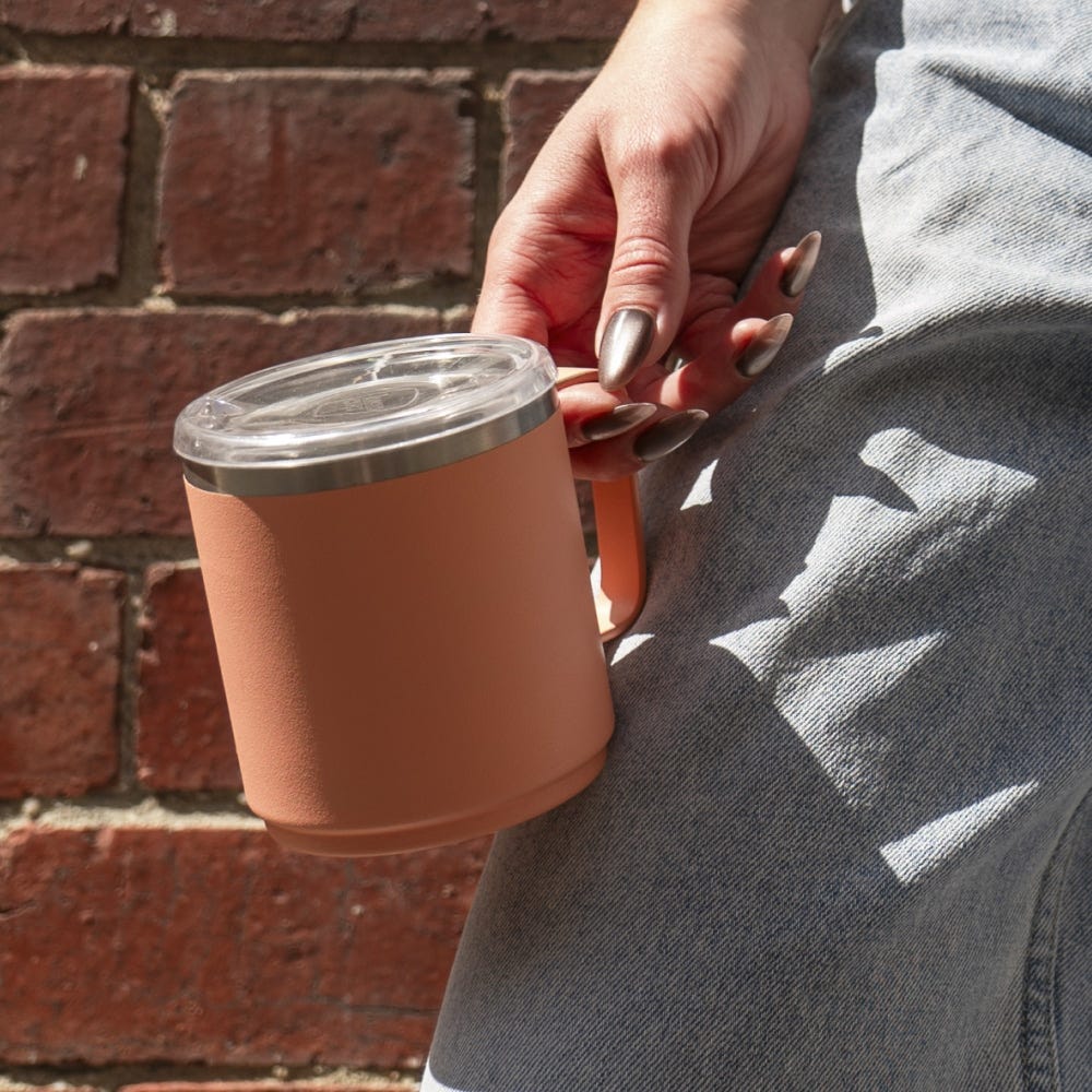 Person holding terracotta-coloured insulated mug with clear lid, wearing grey hoodie against brick wall background
