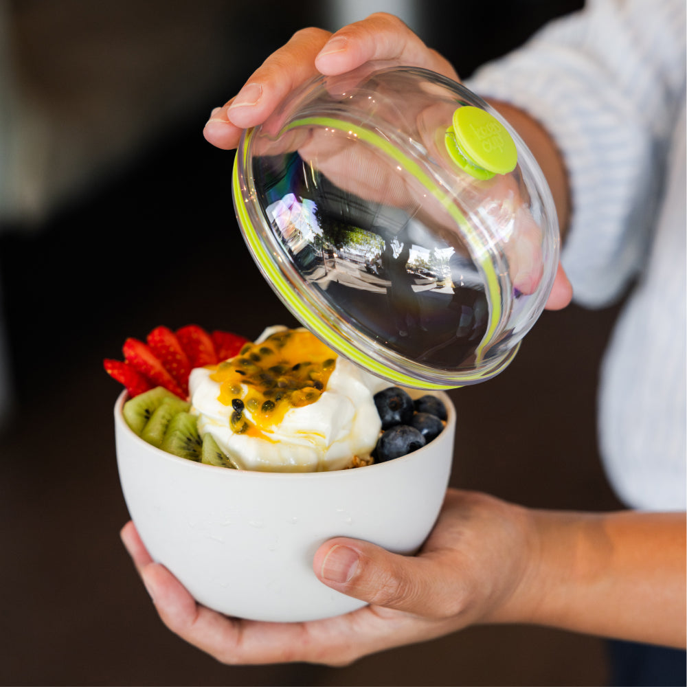 Person holding a white bowl with acai and fruit and a transparent domed green lid.