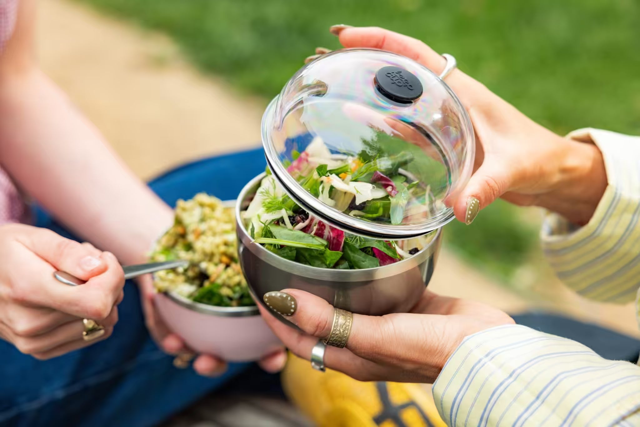 Stainless steel salad bowl with clear glass lid containing fresh mixed greens, held by hands outdoors with picnic setting