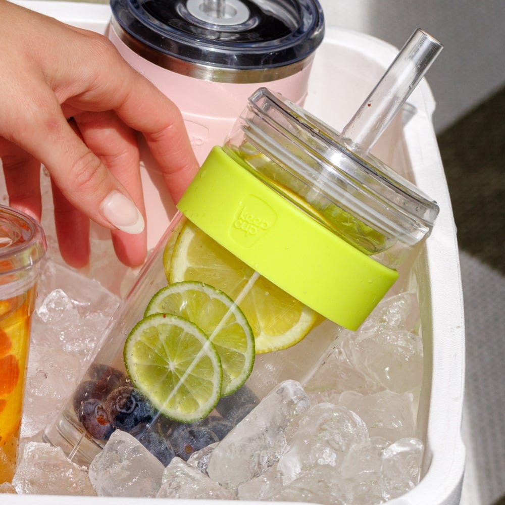 Hand holding a glass cold cup with lemon and lime slices on ice, with a pink keepcup in the background.