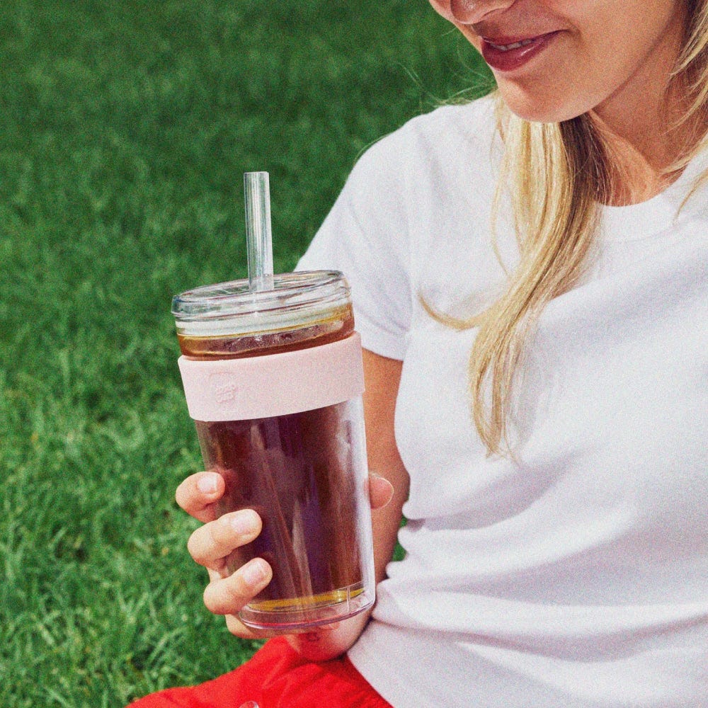 Person holding a glass with a pink band and straw, sitting on grass