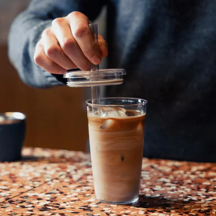 Person using a clear plastic straw to drink iced coffee from a glass on a patterned table.