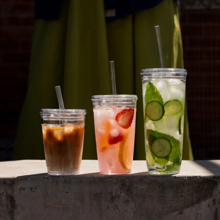Three drinks with straws on a stone surface, one with iced coffee, another with strawberries and lemonade, and the third with cucumbers and mint.
