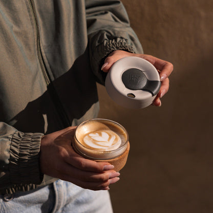 A person in an olive green jacket holds a KeepCup Brew Cork S 8oz in Filter, with the lid removed to reveal a latte featuring a tulip-style milk art design, showcasing the glass cup with cork band and latte lid with soft charcoal plug