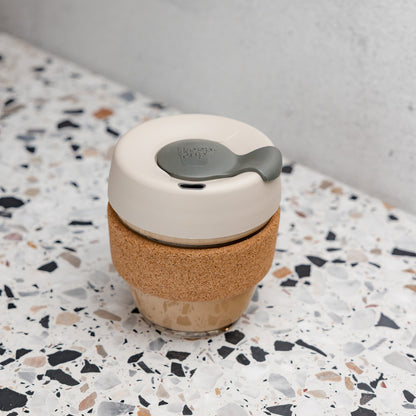 KeepCup Brew Cork XS 6oz in Filter colour, featuring a latte lid with smoke plug and natural cork band, filled with a milky coffee and placed on a terrazzo surface against a concrete wall.