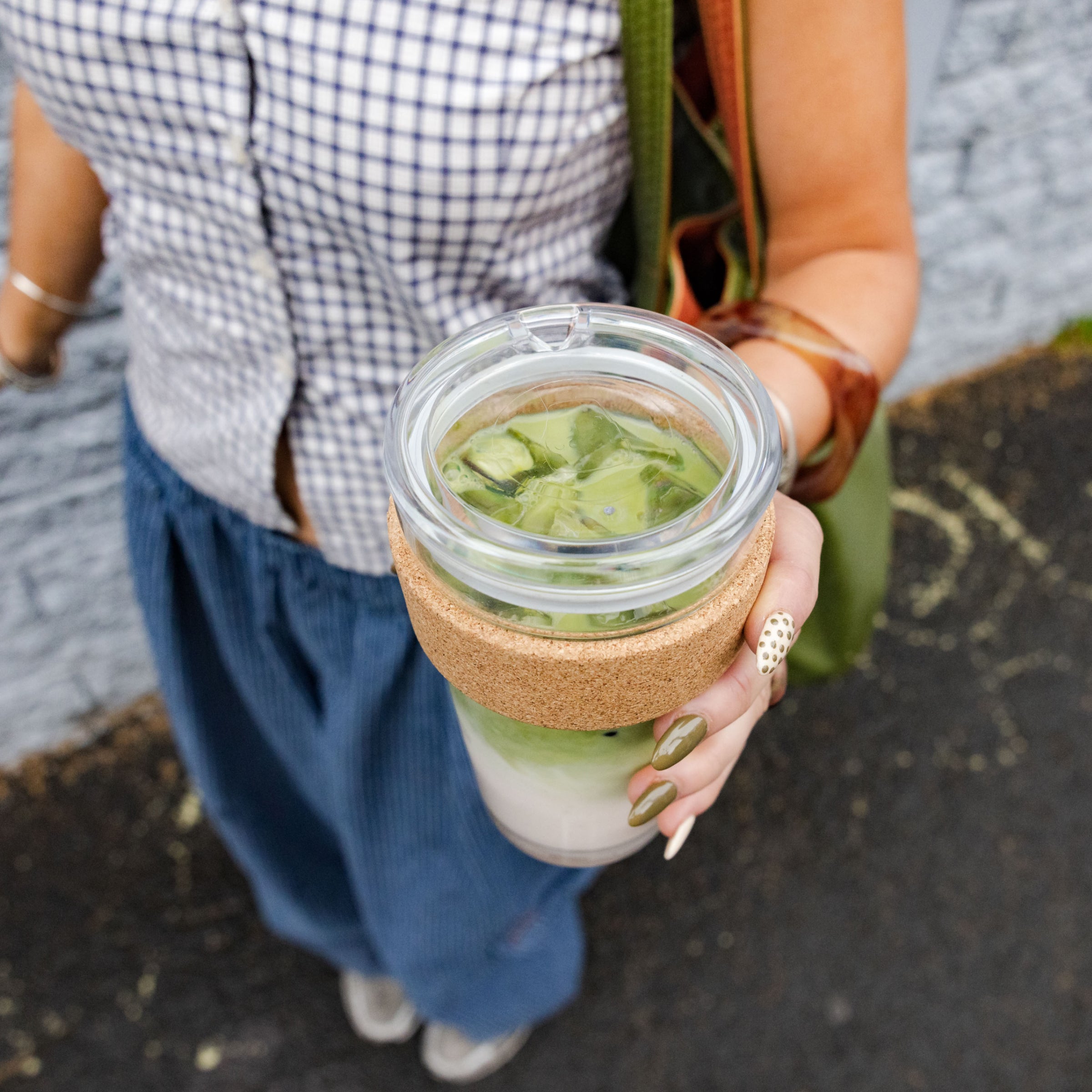 A woman holds a Brew Cork Quicksip L 16oz reusable cup filled with an iced matcha latte, the cork band and clear glass body visible as she stands outdoors wearing a gingham shirt and navy skirt with olive green nail polish.