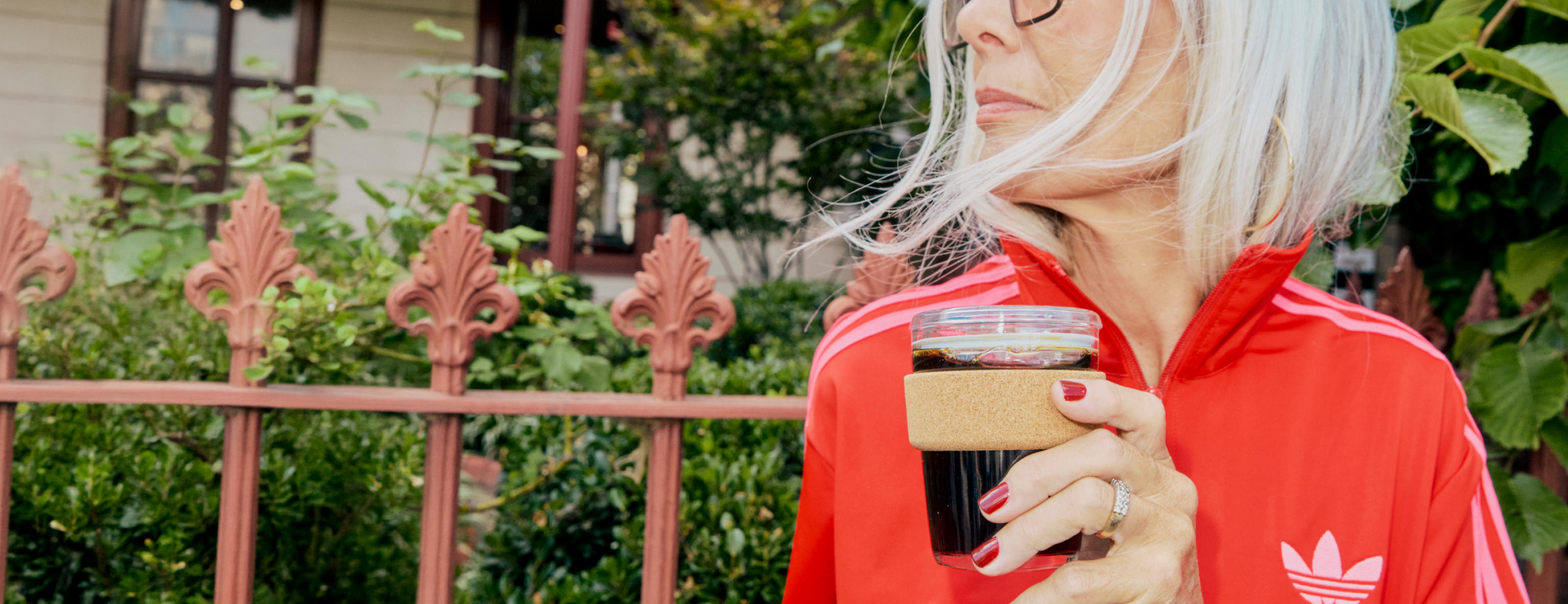 Woman in red Adidas tracksuit holding reusable coffee cup with cork sleeve outdoors by decorative iron fence