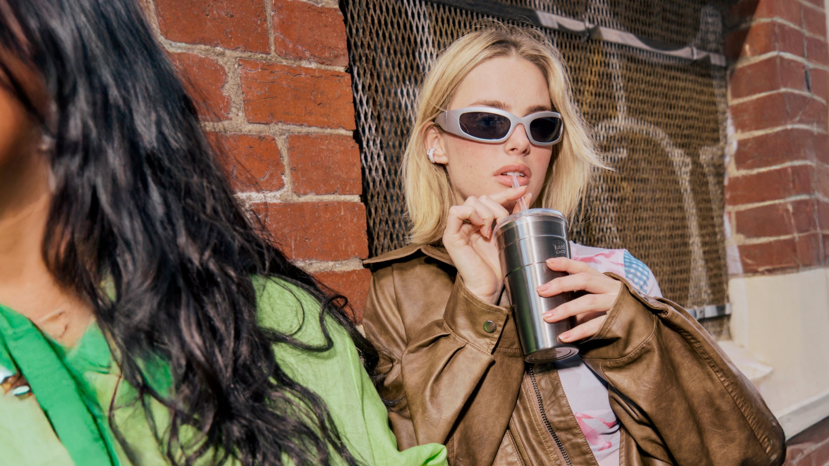 Two women sitting together, one holding a metallic cup, against a brick wall.