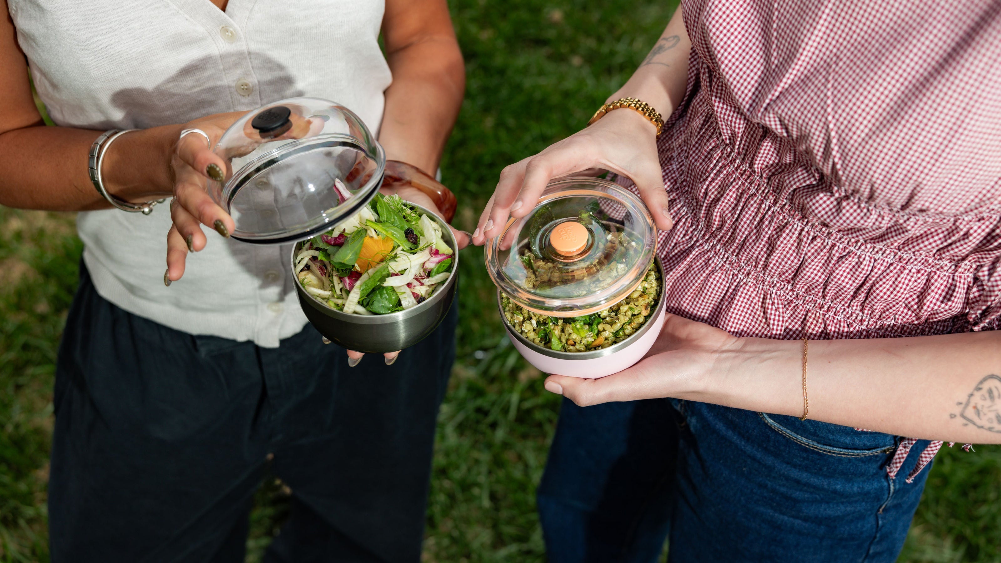 Two people holding containers of salad outdoors on a grassy background