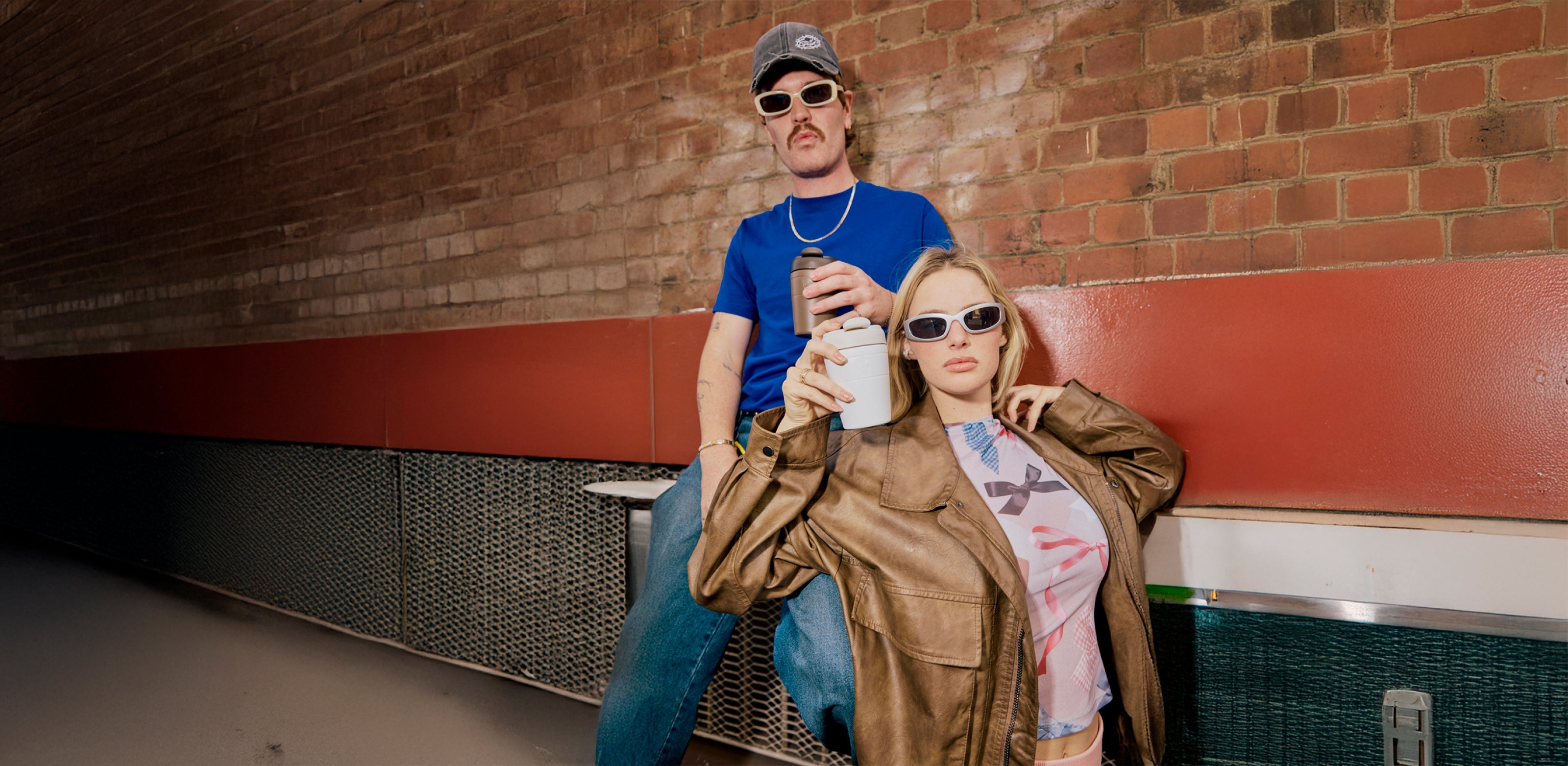 Two people wearing stylish sunglasses in casual outfits - man in blue tee and cap, woman in brown leather jacket holding coffee cup