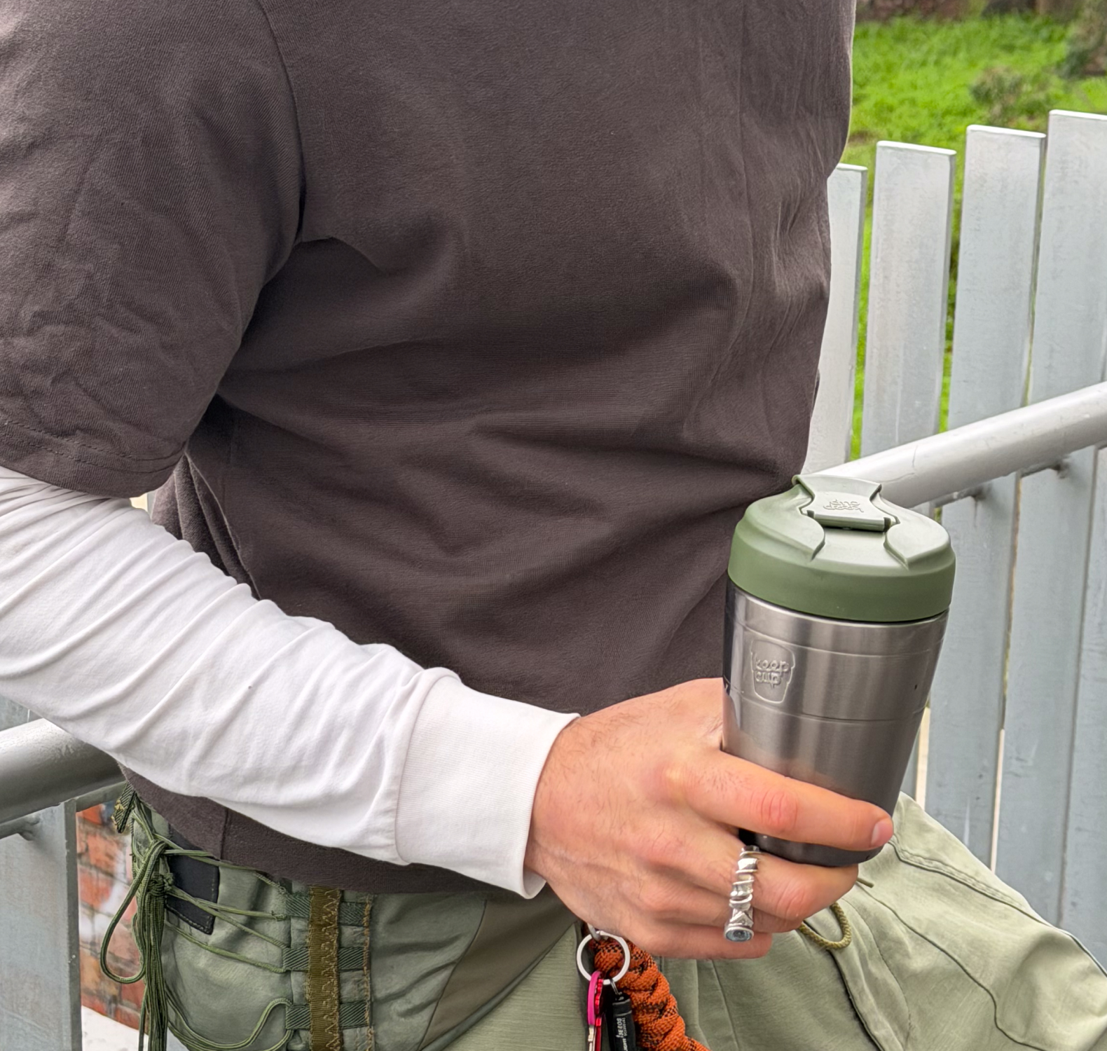 A close-up lifestyle shot of a person holding Commuter insulated travel mug in Nitro Pine. The person is wearing a dark brown shirt, a white long-sleeve undershirt, and olive green pants, standing next to a white metal fence.