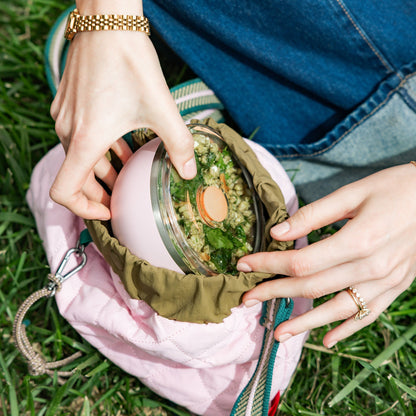Hands removing the flat lid from a Go Bowl Luxe in Pashmak Pink, revealing a grain and greens salad inside, while sitting on grass with the bowl nestled in a quilted pink bag with olive drawstring lining.