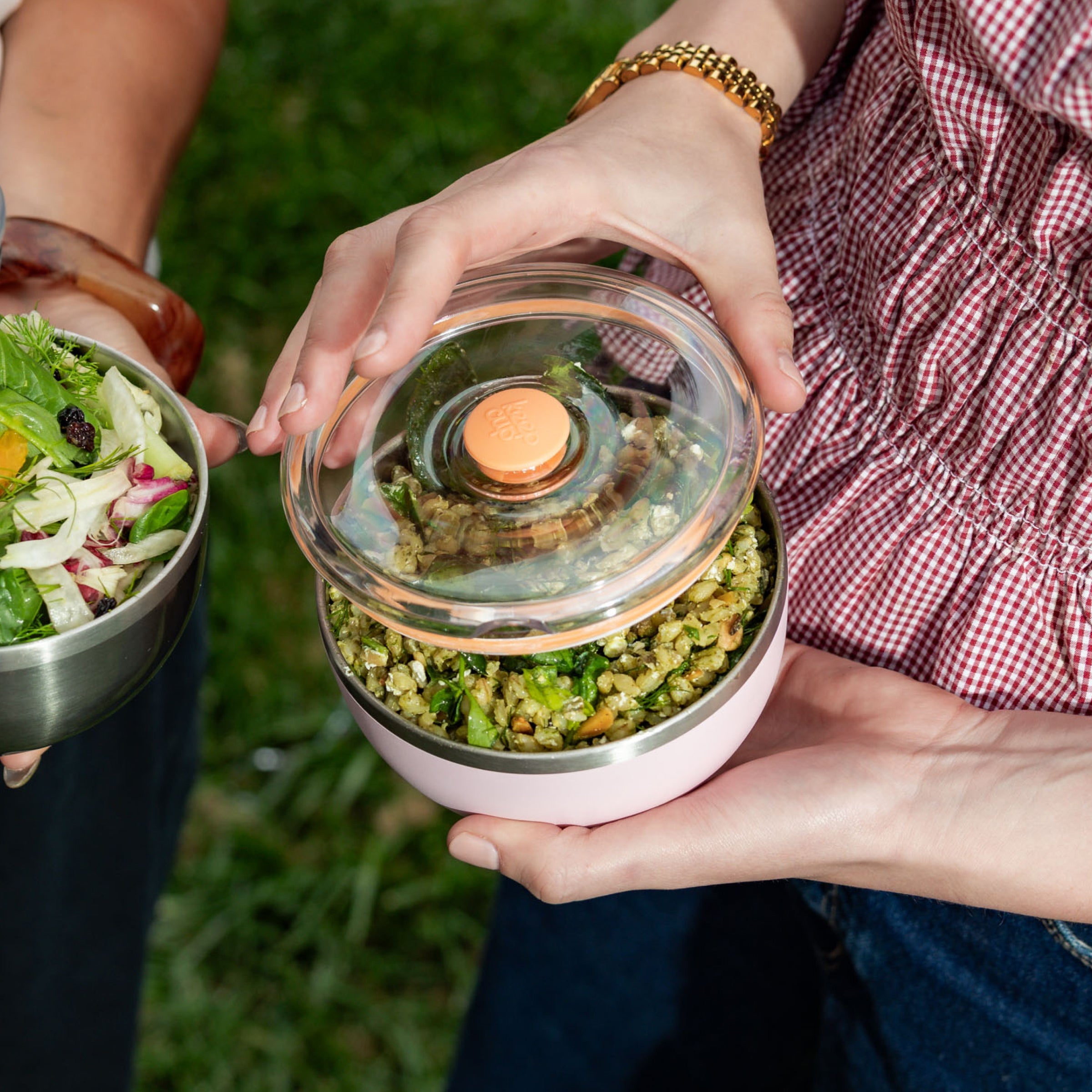 Two people sharing food outdoors, one lifting the flat lid of a KeepCup Go Bowl Luxe in Pashmak Pink filled with a grain salad, while the other holds a stainless steel bowl containing a fresh garden salad.