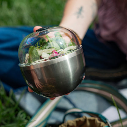 Clear glass salad bowl with stainless steel base containing fresh green leaves and edible flowers, held outdoors