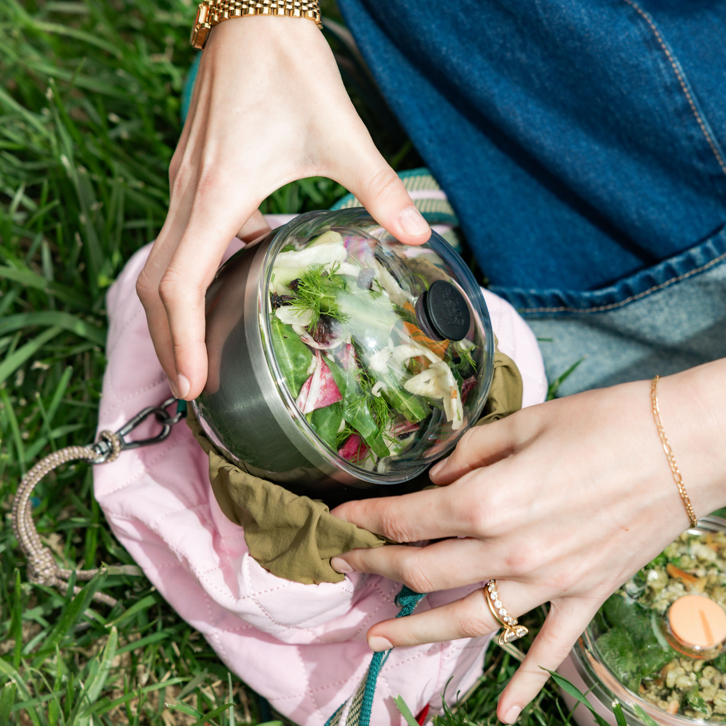 Hands holding stainless steel salad bowl with clear lid containing fresh mixed salad, outdoors on grass with pink bag