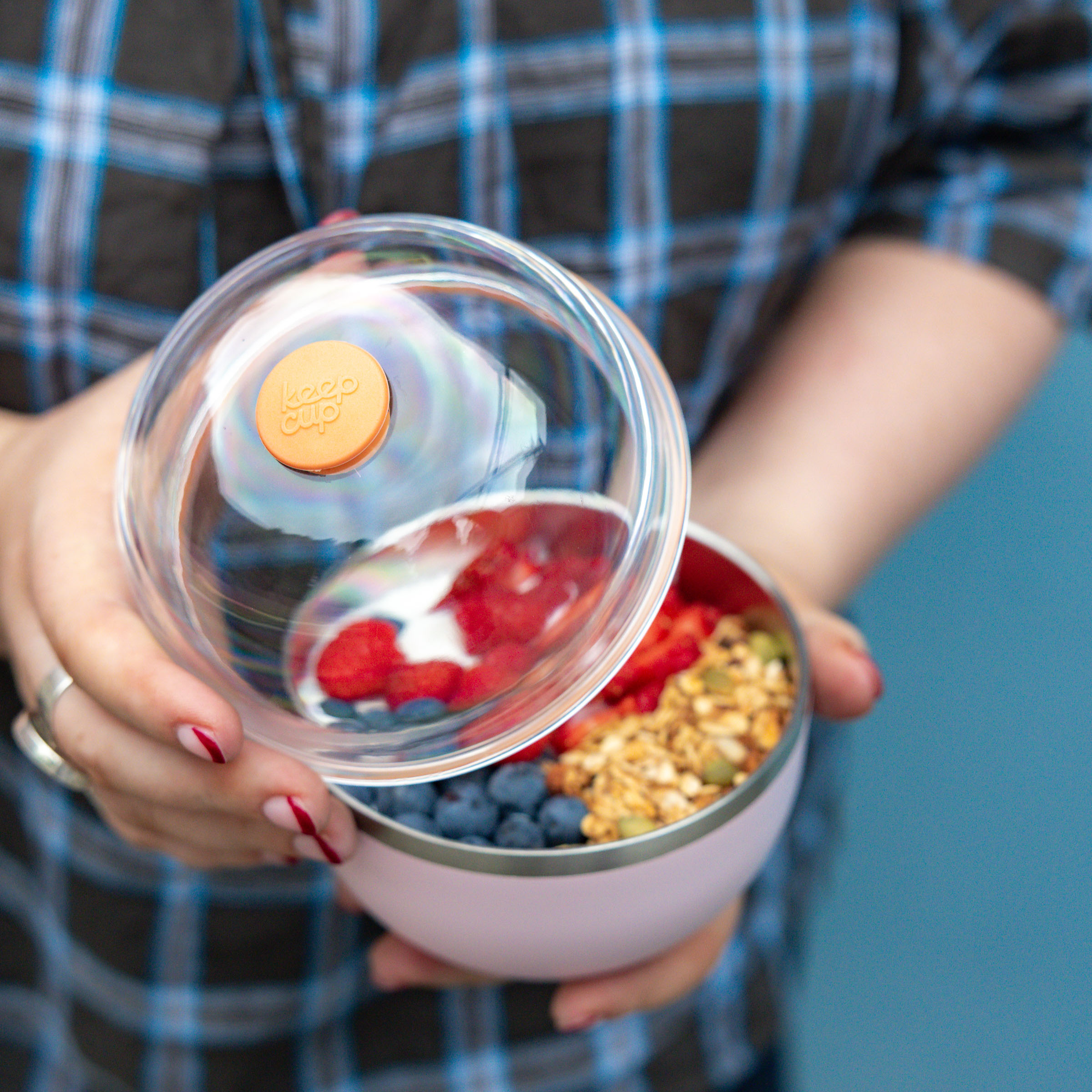 A person in a plaid shirt holds the KeepCup Go High Go Low in Pashmak Pink, lifting the transparent domed lid to reveal a colourful breakfast bowl filled with blueberries, strawberries, granola and yoghurt inside the reusable container.