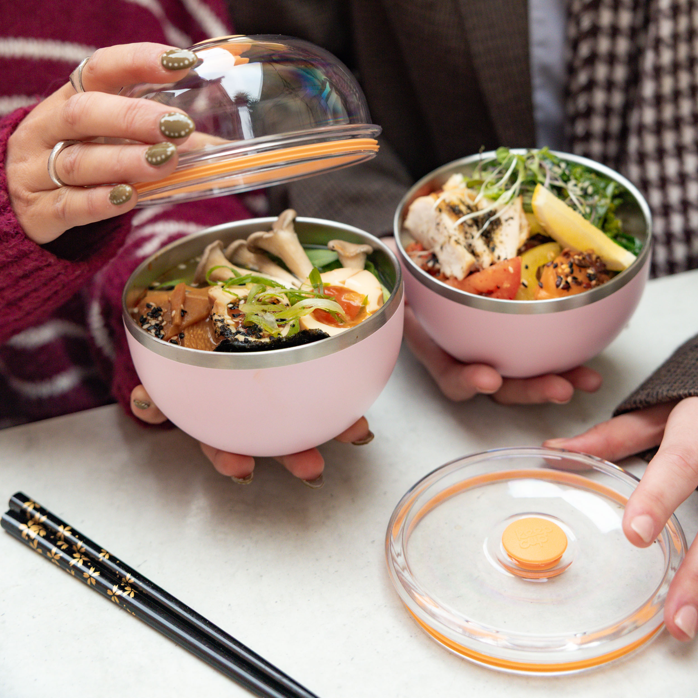 Two people holding KeepCup Go High Go Low bowls in Pashmak Pink filled with Asian-inspired meals featuring mushrooms, tofu, chicken and vegetables, with one person lifting the clear domed lid to reveal the stainless steel interior, chopsticks resting on the table beside them.