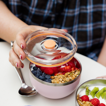 A hand with red nail polish lifts the clear plastic lid off a KeepCup Go High Go Low in Pashmak Pink, revealing a breakfast bowl filled with blueberries, raspberries, strawberries and granola, with a metal spoon resting against the cup on a white table.