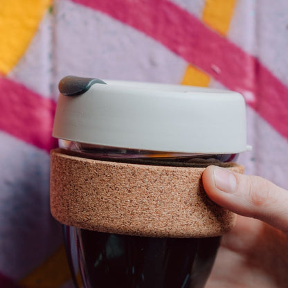 A hand holds a KeepCup Brew Cork XS 6oz in Filter, showing the glass cup filled with dark coffee, natural cork band grip, and latte lid with smoke plug, against a colourful striped background.