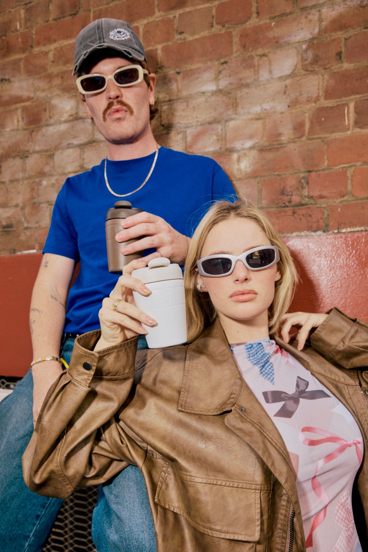 Two people sitting on a couch against a brick wall, holding drinks.