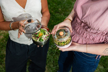 Two people outdoors holding glass lunch containers with lids - one black with salad, one pink with green food