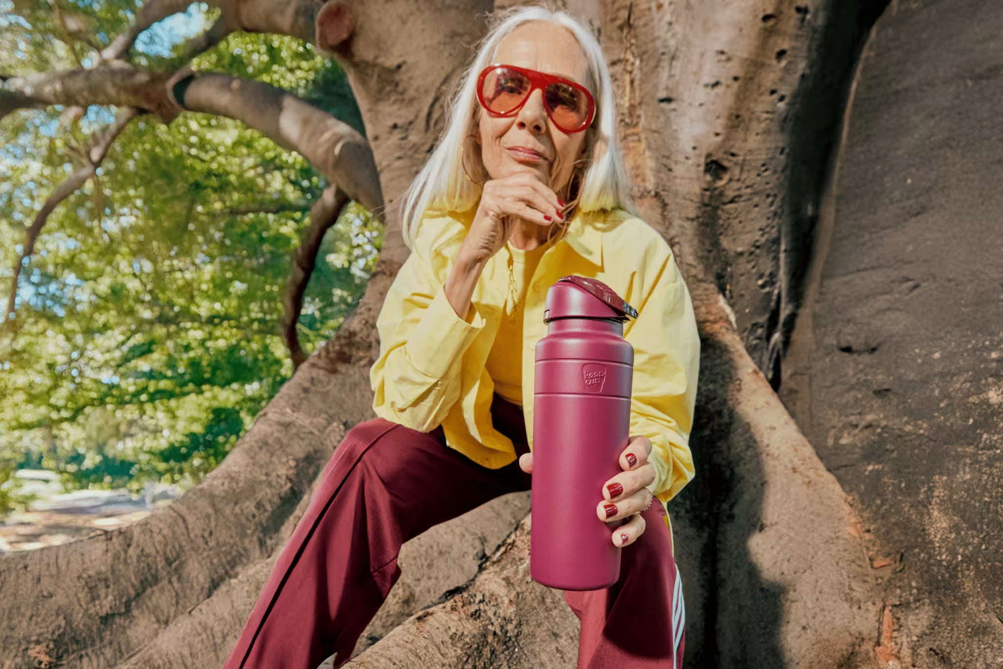 Stylish woman with burgundy insulated water bottle sitting under tree, wearing yellow shirt and red sunglasses in natural outdoor setting