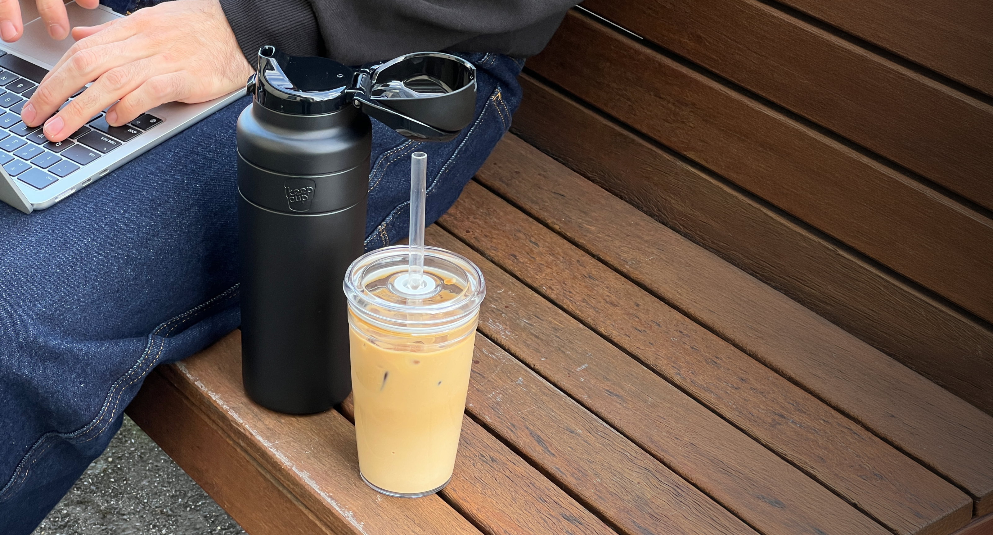 Black insulated water bottle with clip lid next to iced coffee in clear tumbler with straw on wooden bench