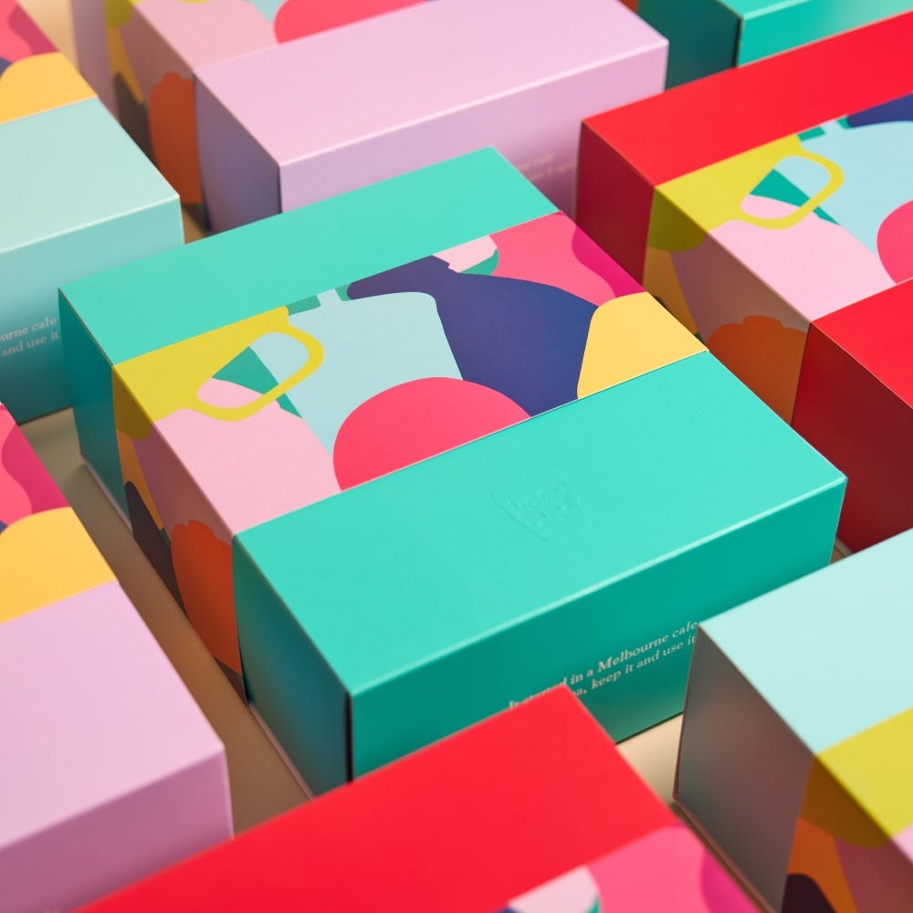 Colourful KeepCup gift boxes arranged in a grid pattern, featuring vibrant packaging in teal, pink, red and yellow with abstract illustrations of reusable cups, including the Just Essentials Pashmak Pink range designed in a Melbourne café.